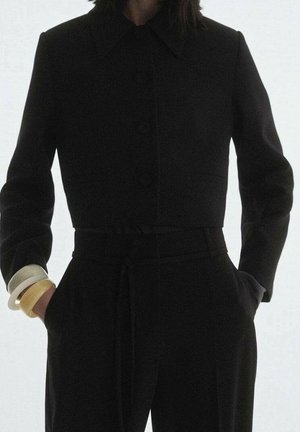 Femme portant une veste noire cintrée et courte avec des boutons, un pantalon noir taille haute avec une ceinture, et trois bracelets sur le poignet gauche.