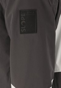 Veste de sport noir et blanc présentant une poche texturée noire avec le texte "SLOPE STYLE" et un design en tissu lisse et élégant.