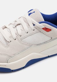 Gros plan sur une chaussure de sport blanche et bleue avec détails en daim et mesh, lacets blancs et accent rouge sur les œillets.