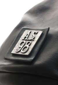 Sac en cuir noir avec une étiquette en métal rectangulaire texturée, embossée du message "A.S. 98." Surface lisse avec un éclat subtil, design minimaliste.