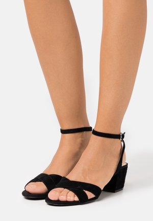 Sandales en daim noir avec un talon épais, dotées de brides croisées sur les orteils et d'une bride à la cheville avec une boucle argentée pour un ajustement sécurisé.