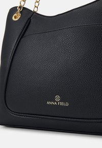 Borsa nera in ecopelle texturizzata, con logo dorato e dettagli in catena, caratterizzata da un design curvo e bordi lisci.
