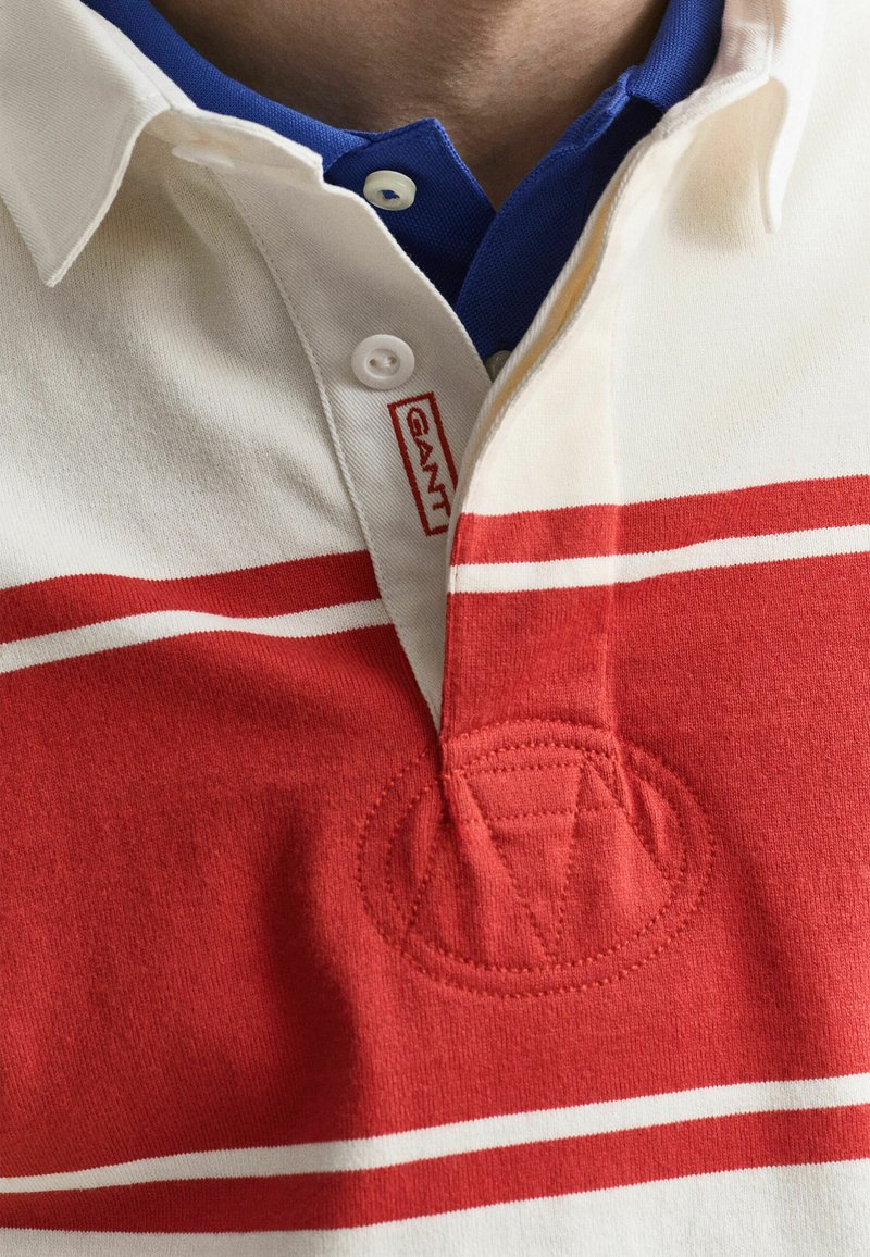 Weiß-rot gestreiftes Polohemd mit blauem Kragen darunter, mit kleinem rotem GANT-Logo nahe den Knöpfen.