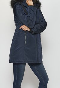 Chaqueta acolchada azul marino con capucha de piel sintética, cremallera completa al frente y bolsillos laterales. Fabricada con un material brillante, con un dobladillo ligeramente curvado.