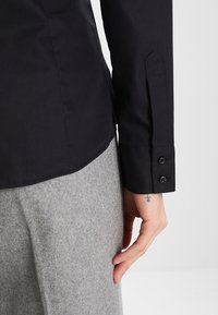 Camicia nera di cotone con polsini abbottonati, texture liscia e un design aderente visibile lungo il lato. Tessuto grigio è visibile sotto.