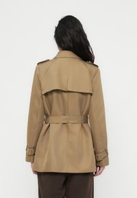 Μπεζ καμπαρντίνα με ζωνάκι στη μέση, επιρράματα στους ώμους και χαρακτηριστικό πίσω μέρος που μοιάζει με κάπα. Λείο υλικό, μινιμαλιστικός σχεδιασμός.