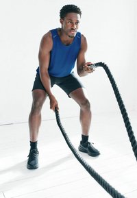 Czarna lina treningowa trzymana przez osobę ubraną w niebieski sportowy tank top i czarne szorty, stojącą na jasnej drewnianej podłodze.