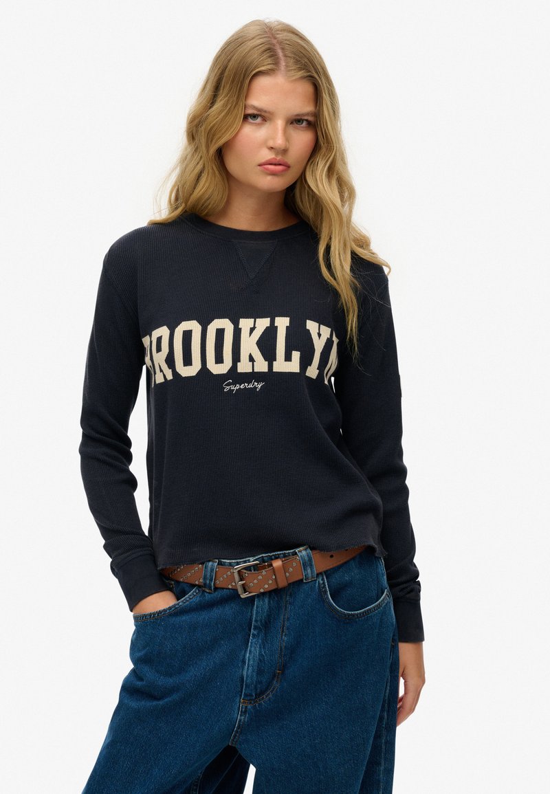 Marineblauwe sweatshirt met lange mouwen met "BROOKLYN" in crèmekleurige tekst; ribbelstructuur; gecombineerd met hoge taille blauwe jeans en een bruine gespbelts.