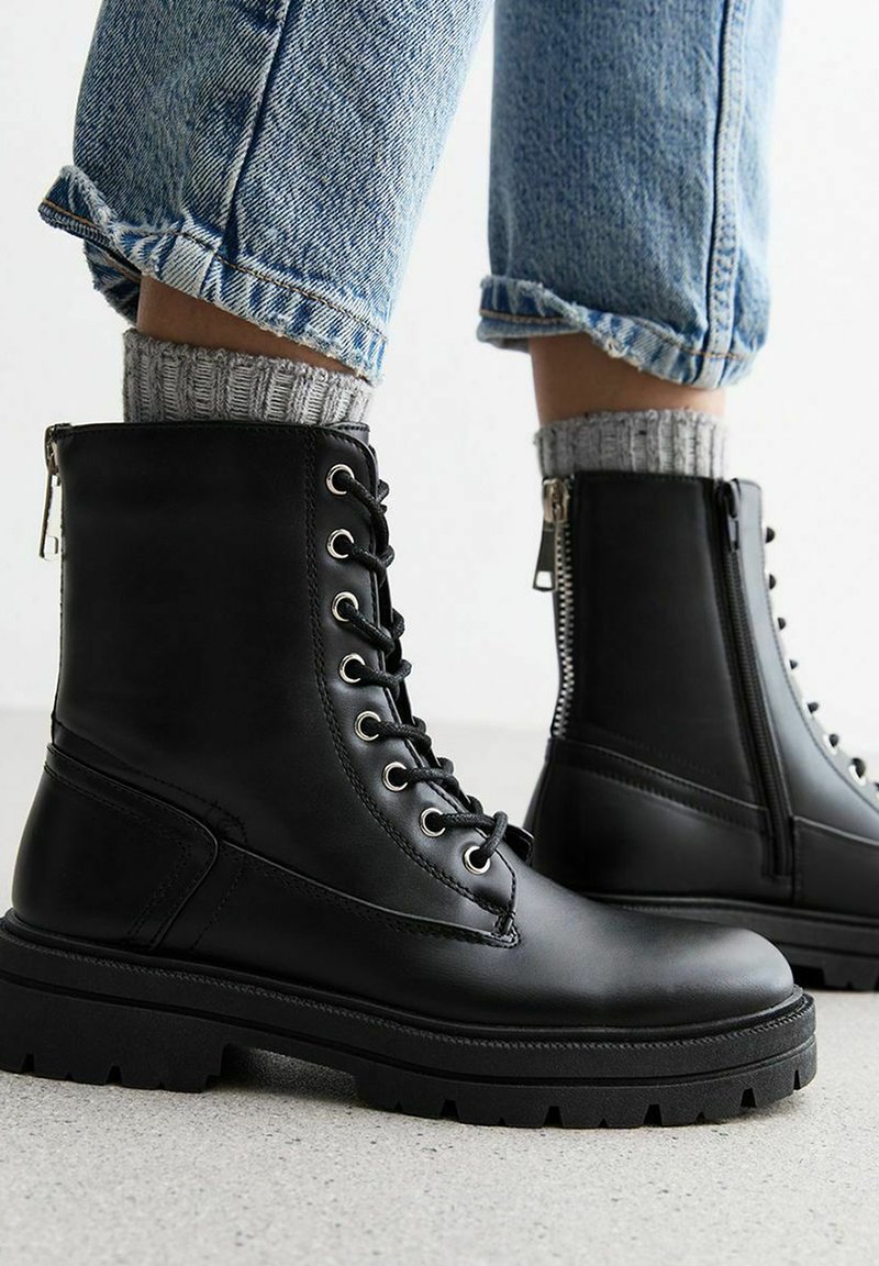 Stivaletti neri in pelle liscia con occhielli in metallo, suola spessa e cerniera laterale, indossati con jeans risvoltati e calzini grigi.