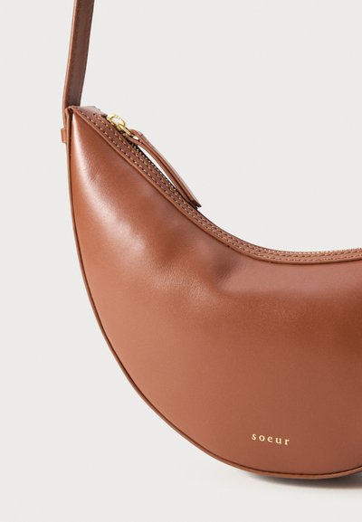 Bolsa de ombro em couro castanho, em forma de crescente, com fecho dourado e pequeno logótipo dourado "soeur" na frente.