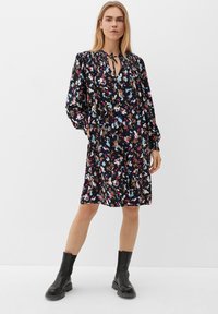 Schwarzes Blumenkleid mit langen Ärmeln, gerafftem Ausschnitt und Bindeband. Verfügt über ein buntes abstraktes Muster und eine lockere Passform. Kombiniert mit schwarzen Stiefeln.
