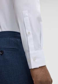 Rechter Arm in einem weißen Hemdsärmel mit geknöpftem Manschette, neben blauen strukturierten Anzughosen mit geknöpftem Gesäßtasche.