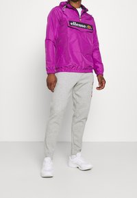 Fioletowa bluza z nylonu z kieszenią z przodu i logo, zestawiona z jasnoszarymi dresami i białymi trampkami. Gładka tekstura, casualowy design.