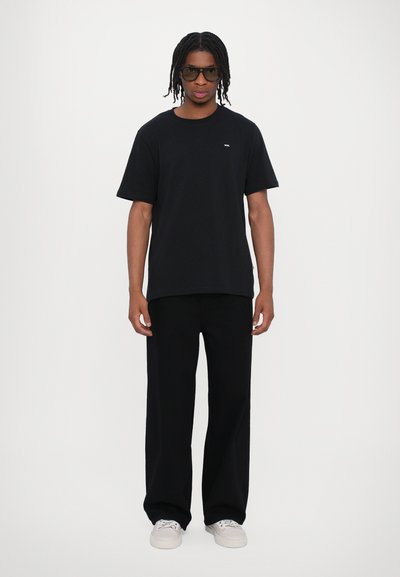 Tricou din bumbac negru cu mâneci scurte, asortat cu pantaloni negri largi și adidași albi. Accesoriile includ ochelari de soare închiși la culoare.