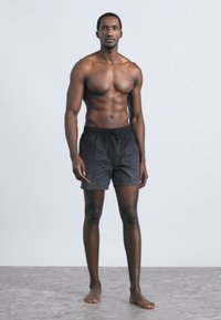 Črne kopalne kratke hlače z nežnim vzorcem, elastičnim pasom in vrvico za zavezovanje, ki jih nosi moški model z golim prsnim košem na enobarvnem ozadju.