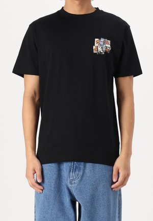 Mand iført sort t-shirt med lille farverigt motiv på venstre bryst og løstsiddende blå jeans, stående mod hvid baggrund.