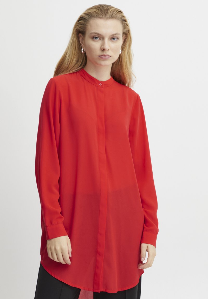 Blusa rossa a maniche lunghe in tessuto leggero e trasparente, con colletto alto, chiusura a bottoni e orlo curvo, indossata con pantaloni neri.