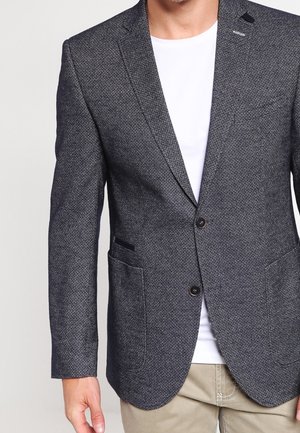Mann trägt einen strukturierten grauen Blazer mit schwarzen Knöpfen über einem weißen T-Shirt und beigen Hosen, steht vor einem schlichten Hintergrund.