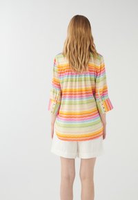 Blus med flerfärgade zigzag-mönster och trekvartsärmar, tillverkad av lätt tyg; fållen är längre bak än fram.