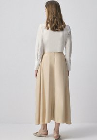 Woman with shoulder-length hair wearing a white long-sleeve blouse, beige midi skirt, and beige low-heeled slingback shoes, standing facing away.