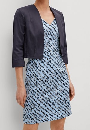 Kvinde iført en lyseblå mønstret kjole med V-hals og en sort cropped blazer med trekvartærmer.