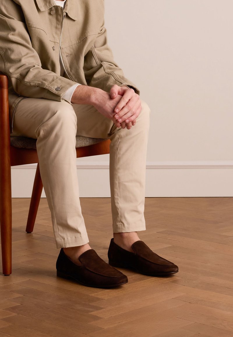 Des chaussures en daim brun foncé à enfiler avec une texture lisse et un design à bout rond, assorties à un pantalon clair et ajusté ainsi qu'un manteau de couleur neutre.