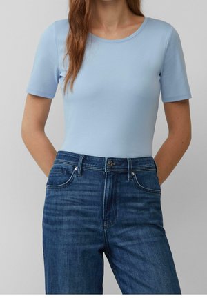 Mujer con camisa de manga corta azul claro metida en unos vaqueros azules oscuros de talle alto, con las manos detrás de la espalda.