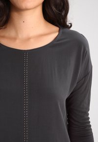 Maglietta grigia scura a maniche lunghe con scollatura rotonda e una fila verticale di borchie decorative lungo la cucitura centrale anteriore. Tessuto morbido.