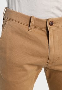 Khaki kalhoty vyrobené z odolné bavlněné tkaniny, s běžným střihem, hnědým zapínáním na knoflík a jemnými švy.