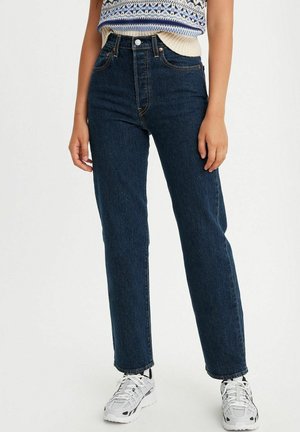 Personne portant un jean droit bleu foncé, des baskets blanches et noires, et un pull sans manches bleu et blanc à motifs.