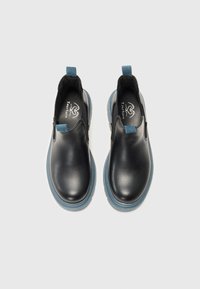 Sapatos pretos de couro tipo slip-on com sola de borracha azul, apresentando uma textura suave e detalhes contrastantes azuis no calcanhar e nas laterais elásticas.