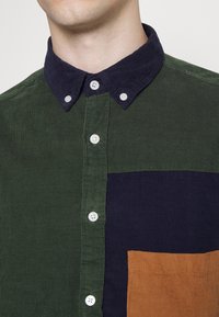 Ribbade skjorta med en mörk marinblå krage, grön kropp och en stor brun och marinblå blockficka, med vita knappar och vertikal textur.