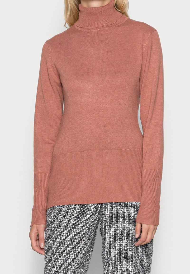 Maglione a collo alto rosa chiaro con maniche lunghe, texture a coste e silhouette aderente, abbinato a pantaloni a fantasia bianco e nero.