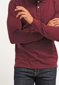 Burgunderfarbendes Langarm-Polohemd mit Kragen, ausgestattet mit drei Knöpfen. Getragen mit dunkelblauen Denim-Jeans, die eine figurbetonte Passform und glatte Textur aufweisen.