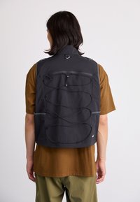 Black vest with adjustable bungee cord detail and two zippered pockets, made from lightweight, textured fabric, worn over a brown shirt.