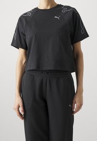 Camiseta negra recortada de manga corta, con diseños abstractos grises en los hombros y un pequeño logo en el pecho, combinada con pantalones a juego.