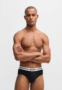 Sorte boxershorts med en hvid elastik, der har "BOSS" logoet og to accenter. Stoffet ser glat ud med en tætsiddende pasform.