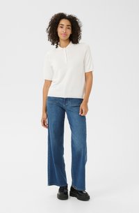 Hvid polo skjorte med korte ærmer, knapper foran, iført blå bredbenede jeans og sorte platform sko. Simpelt design, afslappet stil.