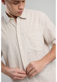 Camicia a maniche corte a righe in beige e bianco. Tessuto leggero con apertura a bottoni davanti, dotata di una tasca sul petto e una texture morbida.