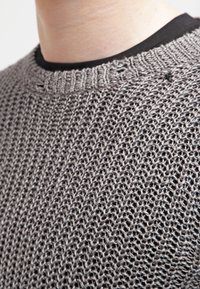 Maglione grigio a maglia con una trama morbida e colletto a coste. La consistenza è aperta e leggera, adatta per essere indossato sopra una camicia nera.