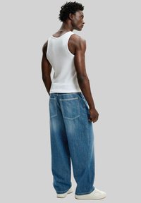 Man i vit linne, vida blå jeans och vita sneakers står med ryggen mot en grå bakgrund.