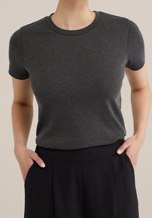 Osoba ubrana w dopasowany ciemnoszary t-shirt z krótkim rękawem i okrągłym dekoltem, wpuszczony w czarne spodnie z wysokim stanem, z rękami w kieszeniach.