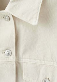 Gros plan du col et des boutons supérieurs d'une veste en denim couleur crème avec des pressions argentées et des coutures visibles.