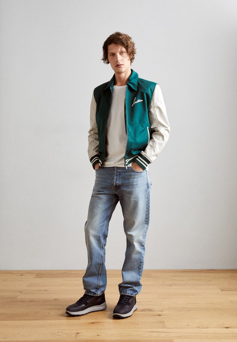 Młody mężczyzna stoi z rękami w kieszeniach, ubrany w teal i biały kurtka typu varsity, beżową koszulę, jasnoniebieskie dżinsy oraz czarne sneakersy na drewnianej podłodze.
