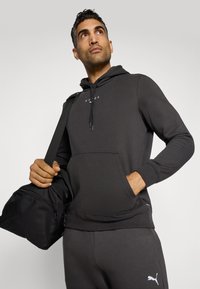 Sweatshirt à capuche noir en matière douce avec poche avant, poignets côtelés et texte logo blanc. Associé à un pantalon foncé assorti et à un sac noir.