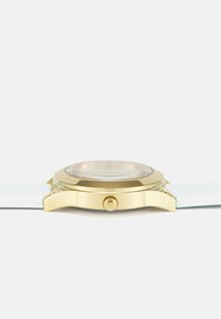 Reloj dorado con caja redonda, correa de silicona blanca y esfera texturizada. Presenta una corona lateral y un detalle brillante en la caja.