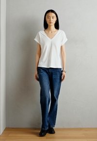 T-shirt blanc à col en v avec des manches retroussées, associé à un jean droit bleu et des chaussures noires. Fond uni, éclairage naturel.