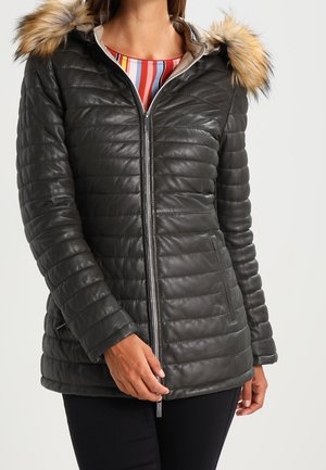 Vrouw draagt een zwarte doorgestikte jas met een capuchon met bontkraag, een ritssluiting aan de voorkant en zakken, gecombineerd met een zwarte broek en een gestreept shirt.