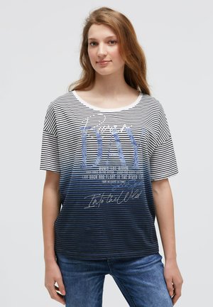 Young woman with long brown hair wearing blue and white striped "Beach Day" graphic t-shirt and blue jeans, standing against a plain background.