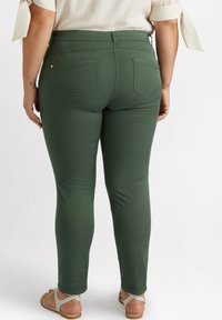 Kvinna som bär olivgröna byxor och beige sandaler, stående med ryggen mot kameran mot en vit bakgrund.